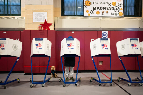 A row of voting booths. Behind them are red gym mats, and above the mats a sign in a window says, Let the March Madness begin!