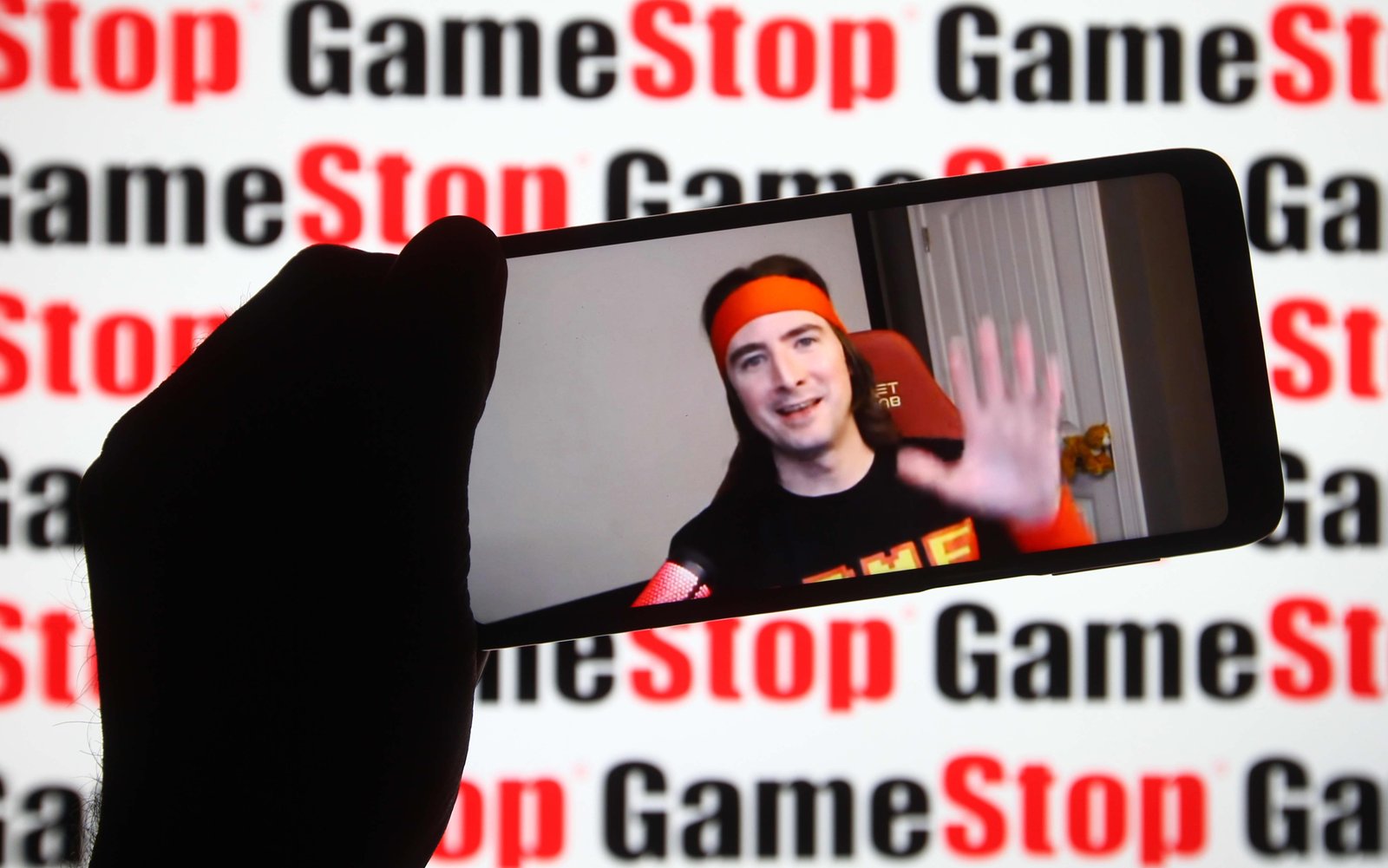 Keith Gill, known on Reddit under the pseudonym DeepFuckingValue and as Roaring Kitty, is seen on a fragment of a youtube video displayed on a smartphone screen in front of GameStop logo.