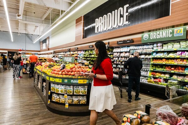 A woman walking inside a grocery store with produce behind her.