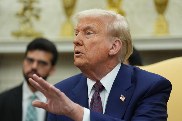 President Trump, seated in a chair, looks to his right while holding up his left hand.