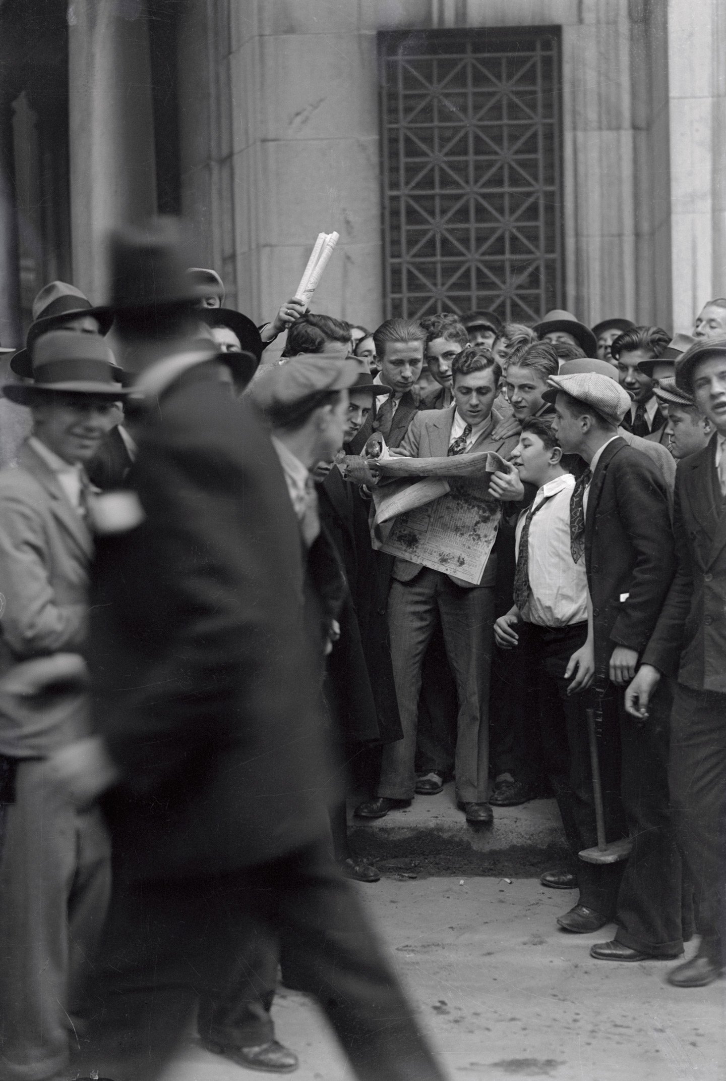 A crowded group of men standing on a street, many wearing hats and suits, gather tightly to read a newspaper. The scene has a vintage or historical feel, with an emphasis on news and community interest. A blurred figure walks by in the foreground.