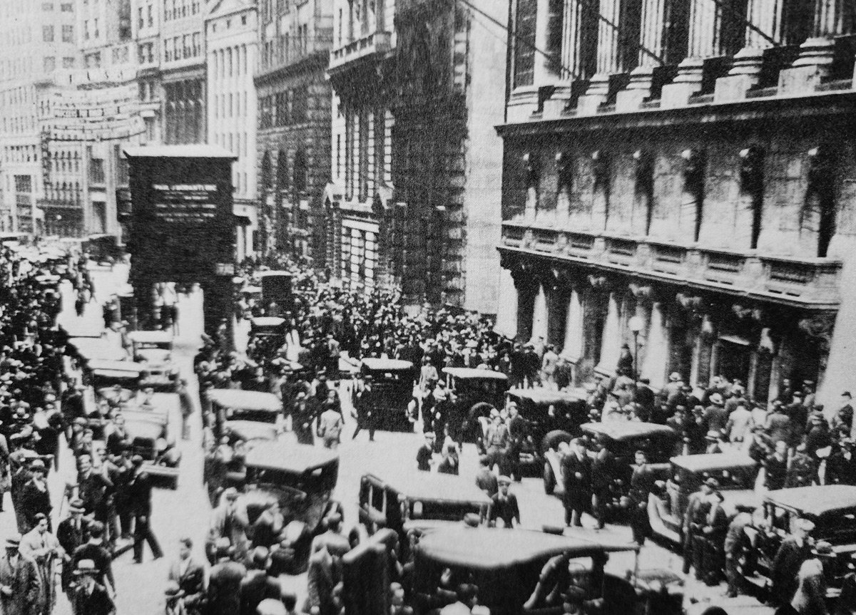 A vintage black-and-white photograph shows a bustling street scene with numerous cars and a large crowd of people surrounded by tall buildings, capturing a lively moment in an early 20th-century city.