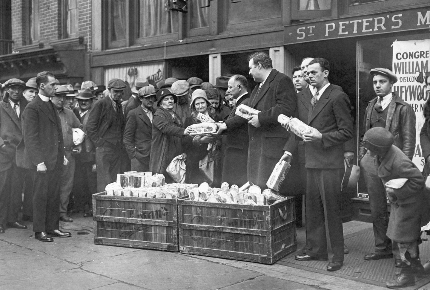 A group of men in suits and overcoats distribute loaves of bread from crates to a line of people. The scene takes place outside a building with a sign that reads 