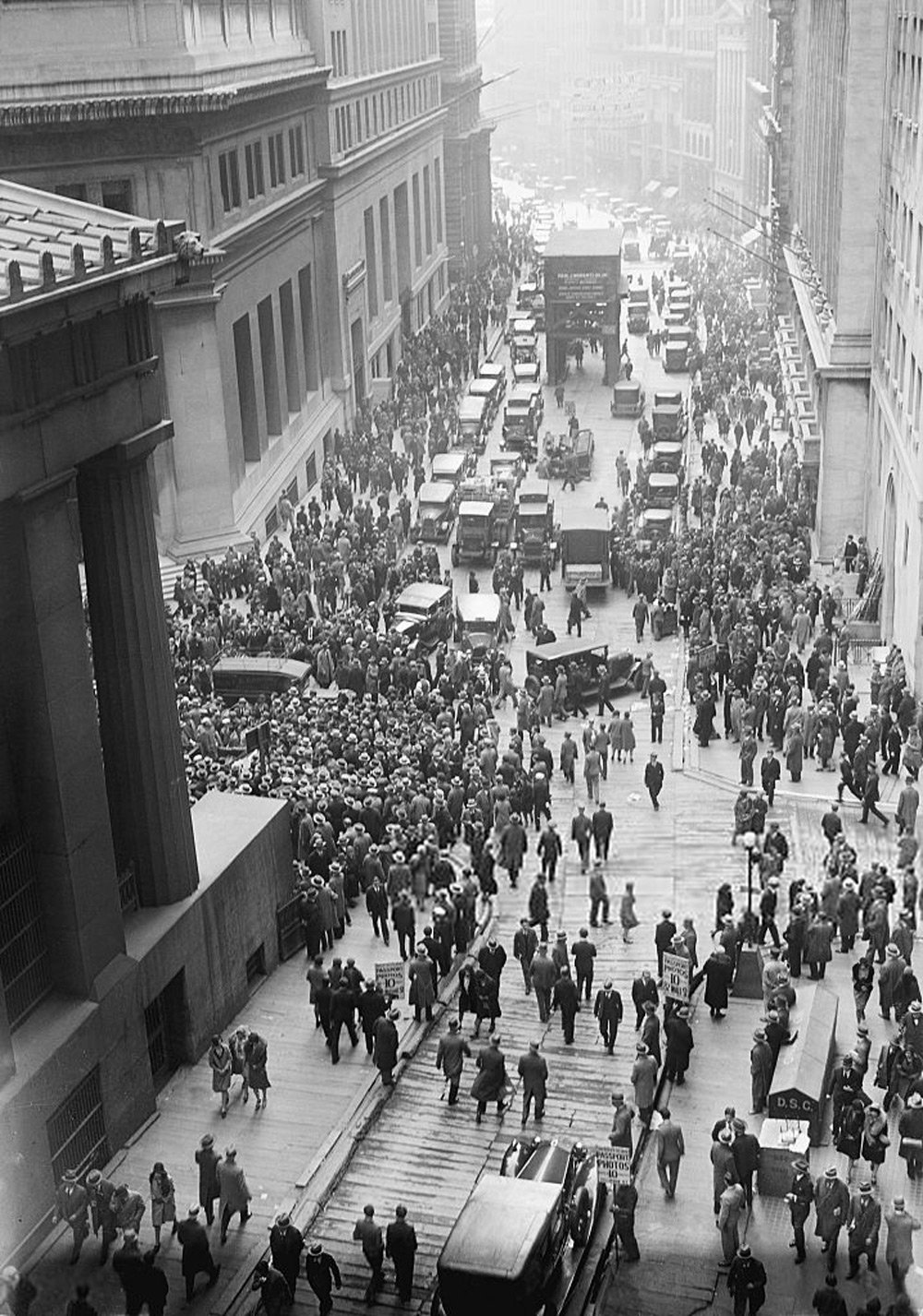 A black-and-white photo of a busy city street in the 1920s or 1930s, filled with people and early 20th-century cars. Large crowds are gathered, and the street is bustling with activity, capturing the energy of an urban setting.