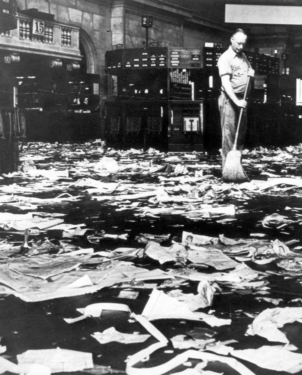 A person sweeps the floor amidst piles of scattered paper in a large, empty room with high ceilings and numbered sections. The scene appears chaotic, with papers covering nearly the entire floor.