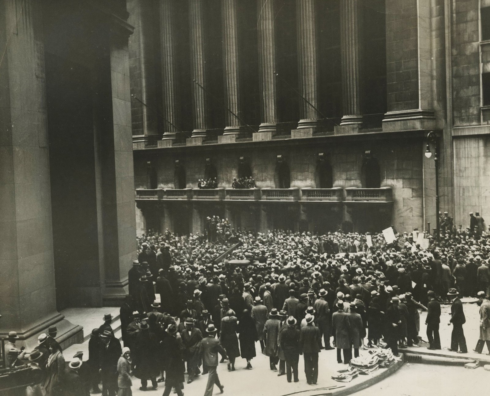 Black and white photo of a large crowd gathered outside a grand building with tall columns, possibly a financial institution. People wear early 20th-century clothing, including hats and overcoats. Some hold signs, suggesting a protest or significant event.