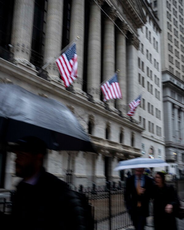 A blurred photo of a colonnaded city building on a rainy day as pedestrians under umbrellas pass by.