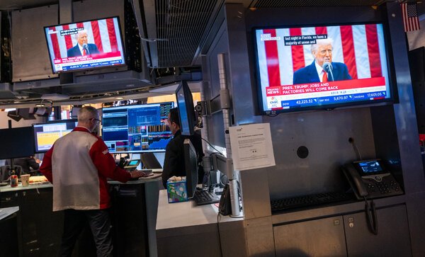 A man standing at a bank of computers, with television screens showing President Trump nearby. 