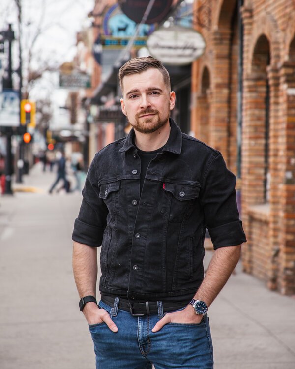 John Kakuk stands on a sidewalk with his hands in his pockets. He is dressed in a black shirt and denim pants.