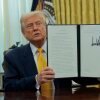 In this photo, President Trump holds up an executive action he signed. He is seated at a desk in the Oval Office and is wearing a dark blue suit and golden-colored tie.
