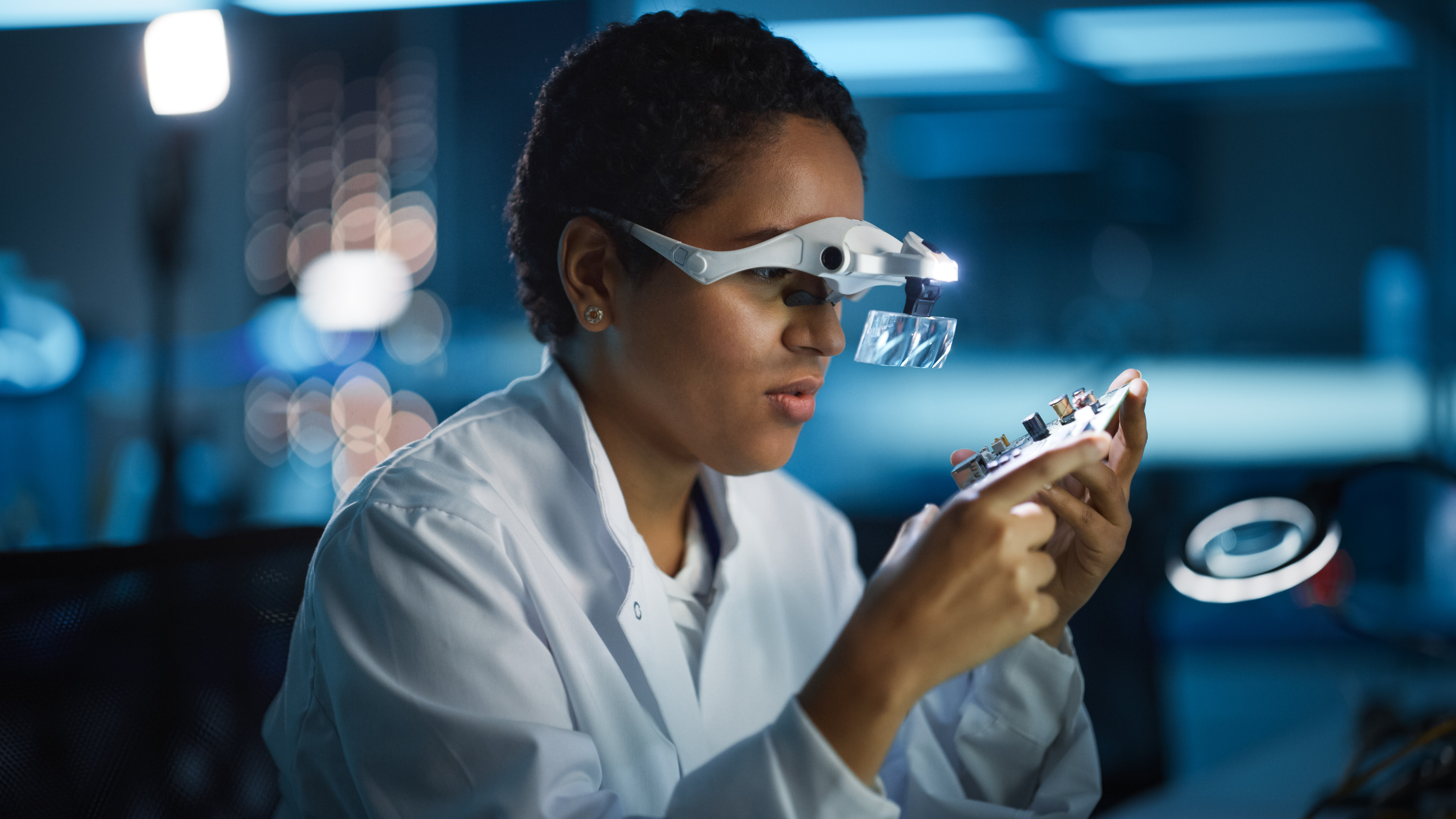 Person in a white lab coat working with a circuit board.