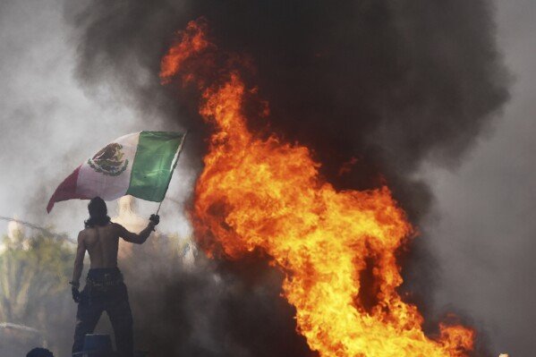 A protester waves a flag as a Waymo self-driving taxi burns near the Metropolitan Detention Center in downtown Los Angeles, June 8, 2025. (AP Photo/Jae C. Hong)