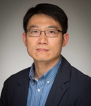 A man with short dark hair and glasses wears a blue and white striped shirt under a dark blazer, looking directly forward with a neutral expression against a gray background.