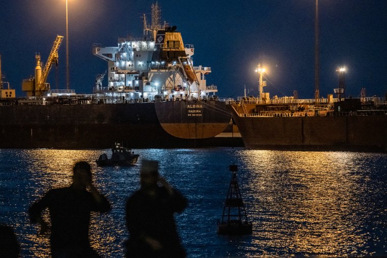 Two men are silhouetted against lights from large tankers floating across the water at night.