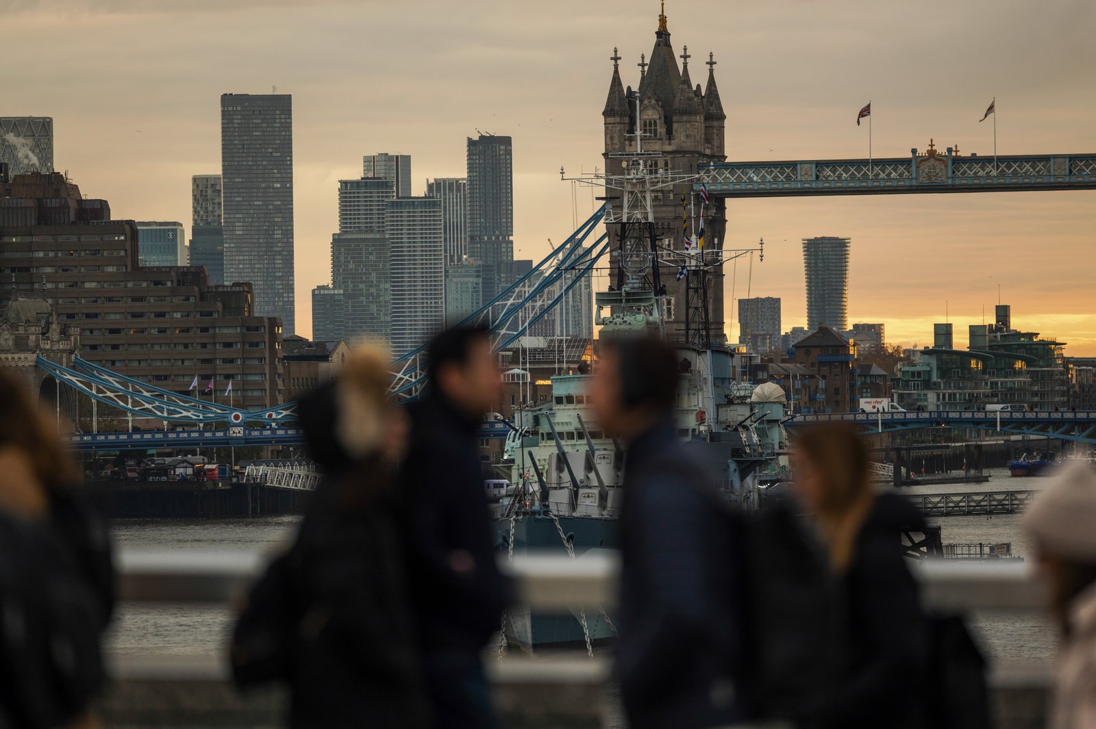 Commuters cross London Bridge with the view to Tower Bridge and the Canary Wharf district in London, UK, on Tuesday, Nov. 18, 2025.