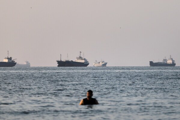 A person stands in shallow water as cargo and commercial vessels are anchored in the Strait of Hormuz off Bandar Abbas, Iran, Monday, June 8, 2026. (Amirhosein Khorgooi/ISNA via AP)