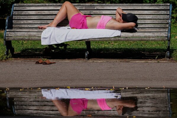A woman is reflected in a puddle as she sunbathes on a bench in Victoria Park, in London, Tuesday, June 23, 2026.(AP Photo/Alberto Pezzali)