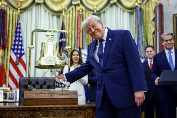 President Trump, in a blue suit, light dress shirt and blue tie, smiles beside a desk as he pulls on a large golden bell.