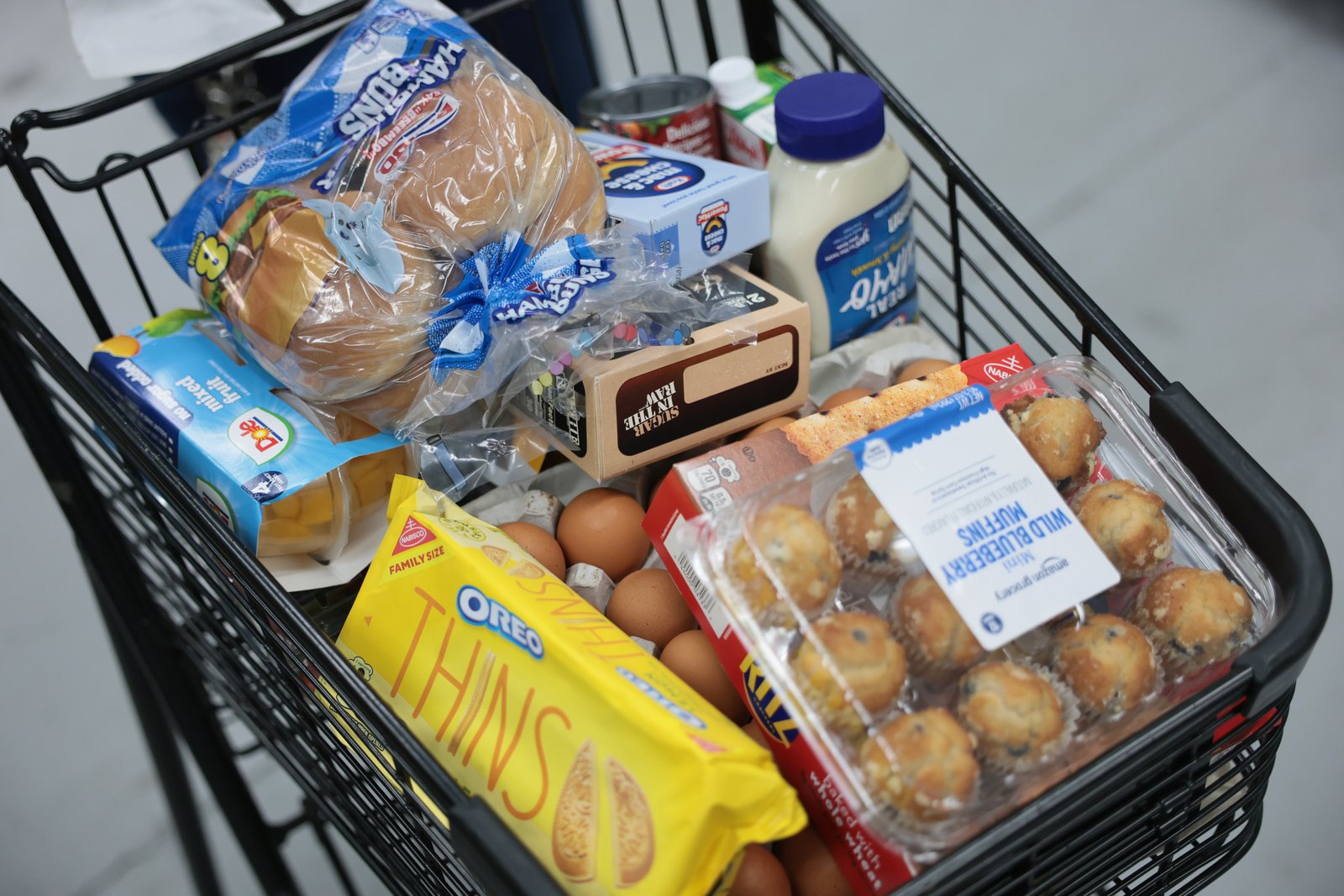 A grocery cart filled with hamburger buns, mixed fruit, sugar, mac and cheese, mayonnaise, eggs, muffins, and Oreo Thins.