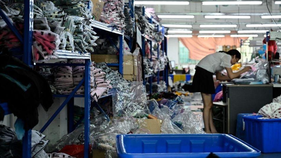 A worker makes clothes at a garment factory that supplies Shein. She is surrounded by piles of clothes stacked on blue shelves.  