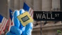 A person's hand, wearing a light blue protective glove, holds a gold-colored CPU (Central Processing Unit) chip with black pins facing the viewer. In the blurred background, a black street sign reads 'WALL ST' with '←22-51' above it. Several American flags are also visible in the soft focus background.