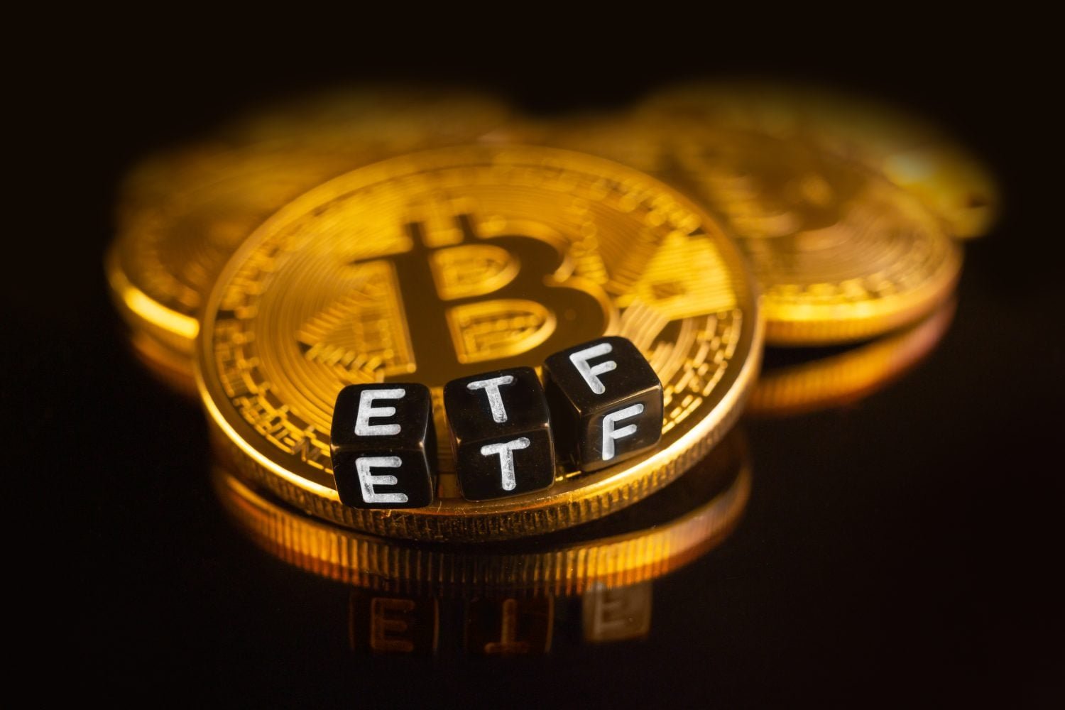 Close-up of several shiny golden Bitcoin coins stacked on a dark, reflective surface. Three black cubic blocks with white letters spelling 'E', 'T', 'F' are placed on the front Bitcoin coin. The background is dark and blurred.