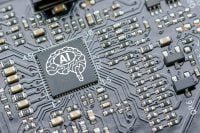 A close-up shot of a dark grey circuit board with a prominent black square microchip. The chip features a white outline of a human brain with the letters 'AI' inside it. Surrounding the chip are many small silver and beige electronic components connected by etched pathways.