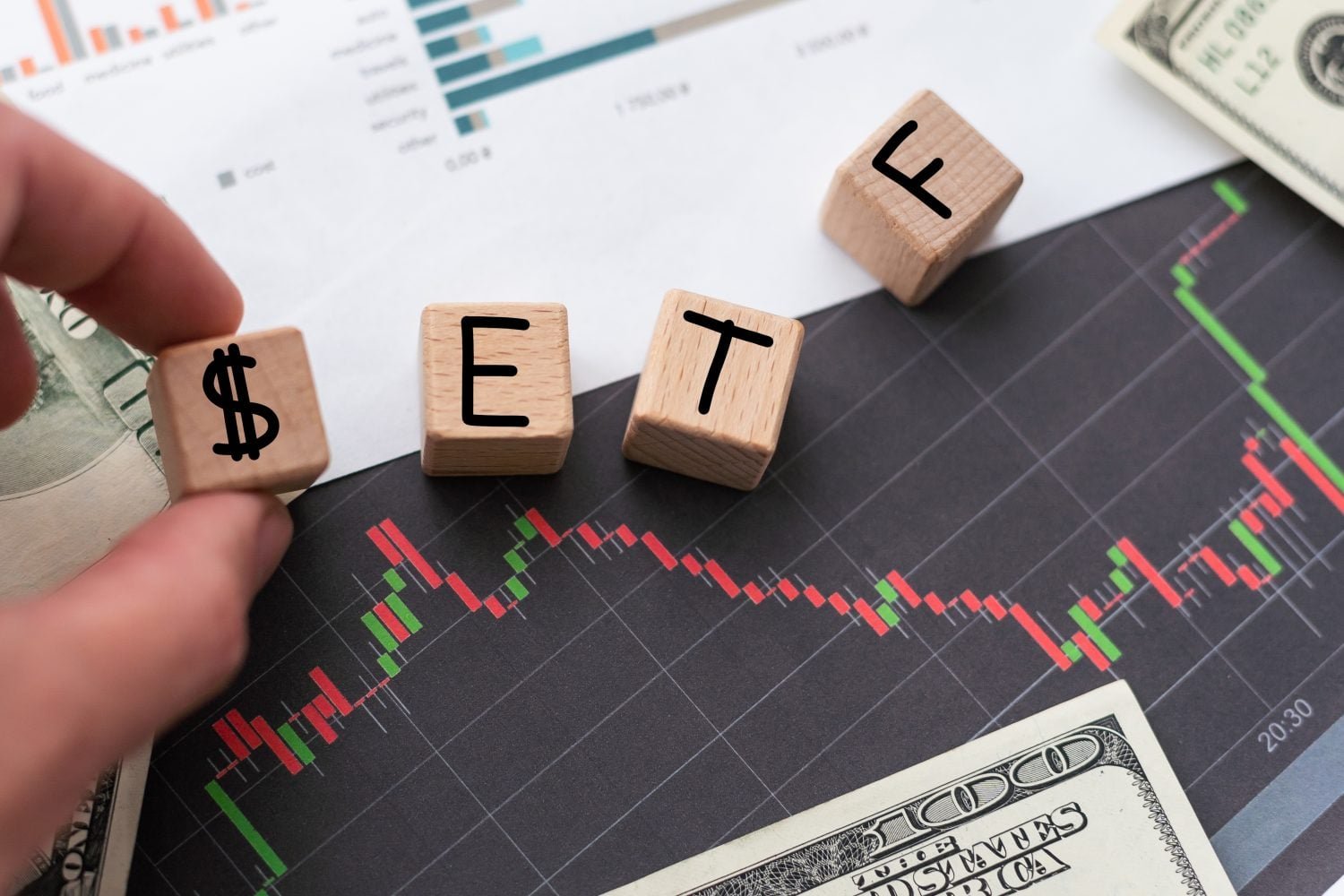 A close-up shot of a hand placing a wooden block with a black dollar sign ($) next to other wooden blocks spelling 'E', 'T', and 'F' on a dark gray financial candlestick chart. The chart shows red and green candles, indicating market fluctuations. Partially visible in the background are US dollar bills and a white paper with bar graphs. The blocks, chart, and money are arranged on a light surface.