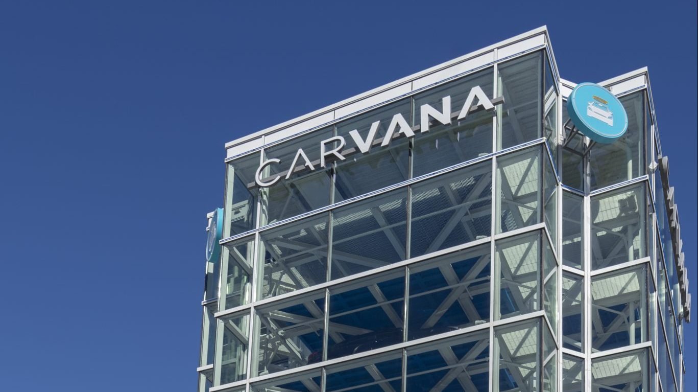 A close-up, low-angle shot of a tall, modern building with a glass facade, prominently displaying the 'CARVANA' logo in large white letters near the top. To the right, a round blue sign with a white car graphic, Carvana's brand icon, is visible. The structure features multiple levels of transparent glass panels and white framework, suggesting a car vending machine. A bright, clear blue sky fills the background.