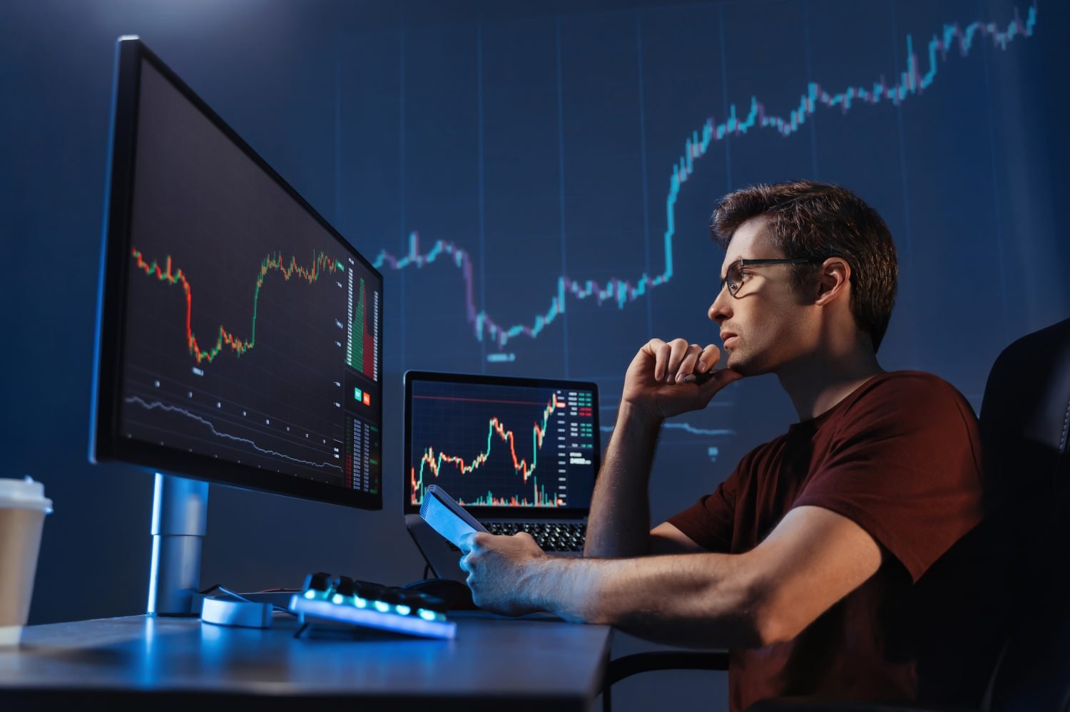 A man with glasses in a red shirt sits at a desk, looking intently at multiple computer monitors displaying financial charts with red and green lines. A laptop also shows similar charts, and a large projection of market data is visible on the wall behind him. He has his hand to his chin in a thoughtful pose, holding a phone or device in his other hand. A backlit keyboard and a coffee cup are also on the desk.